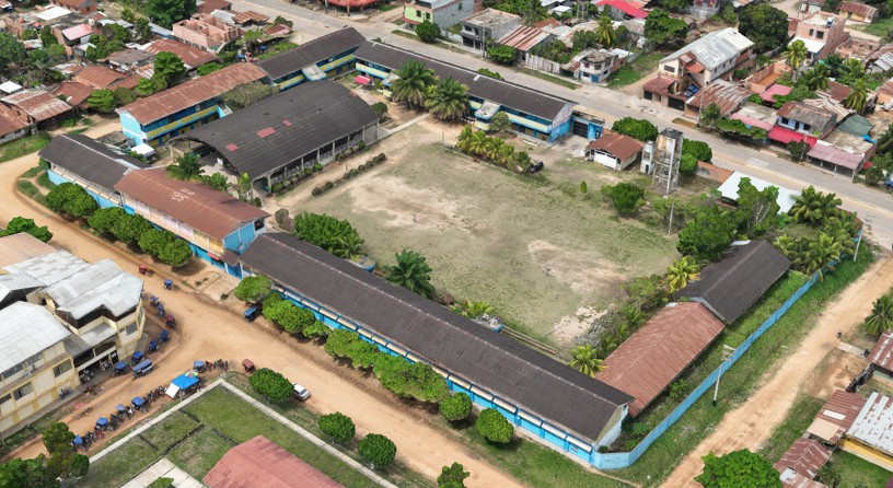 Vista aérea del local escolar: pabellones de techo oscuro y muros celestes alrededor de un amplio campo central.
